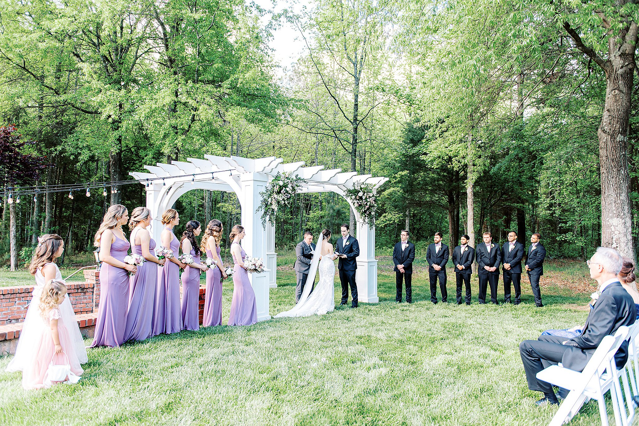 A wedding ceremony with large wedding parties takes place under a white gazebo and tall trees