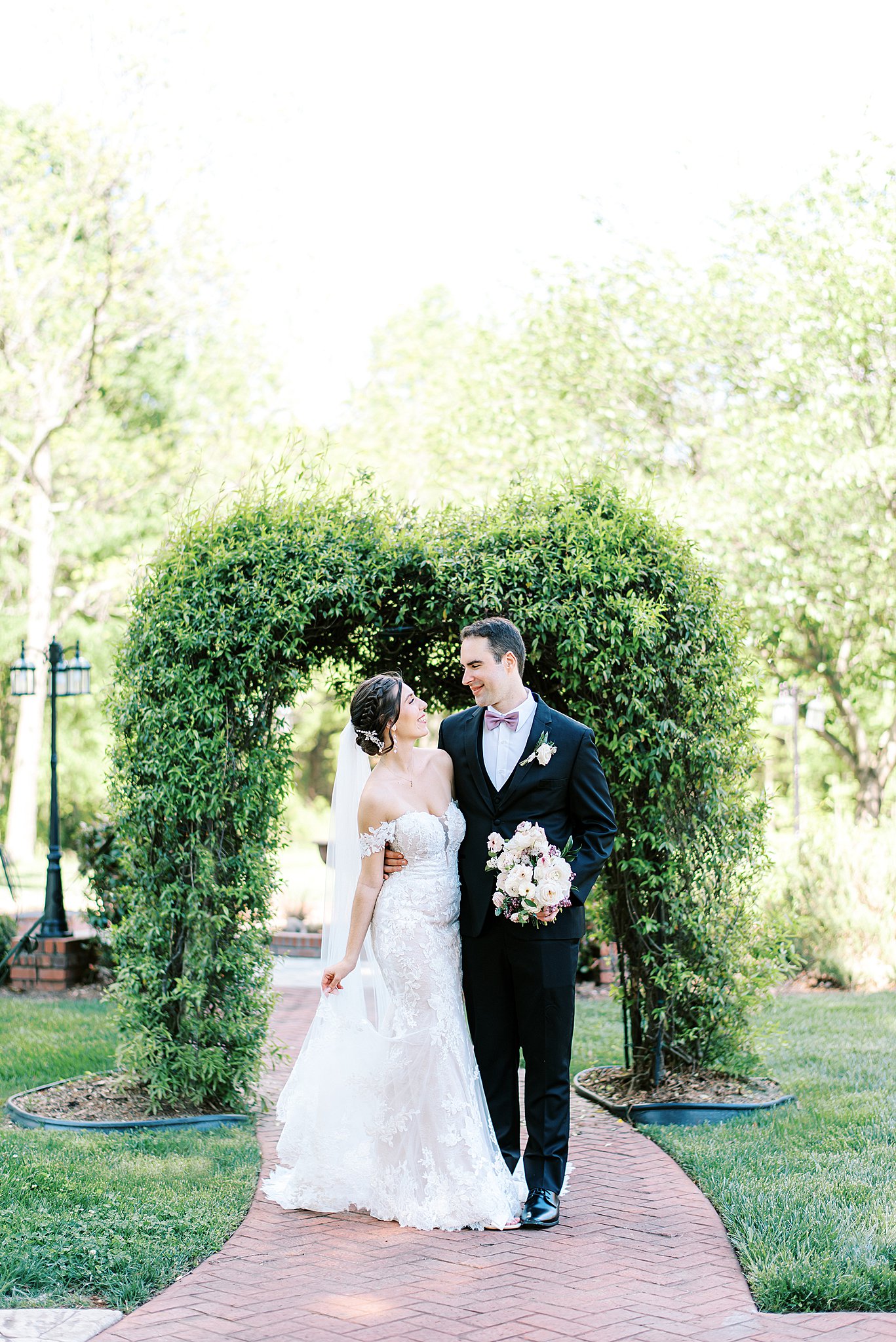 Newlyweds walk through a garden arch in a lace dress and black suit Brawley Estate