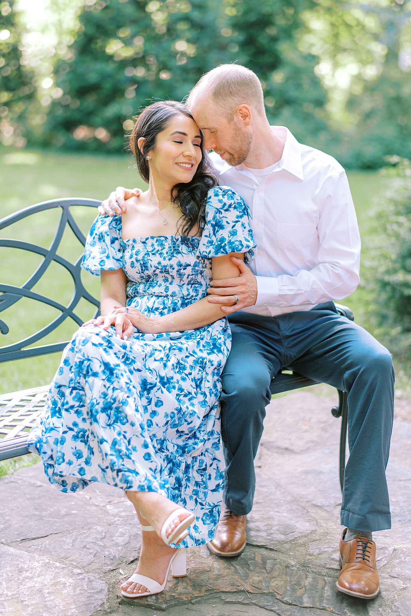 A bride sits on a bench leaning into her husband in a garden while wearing a blue floral dress