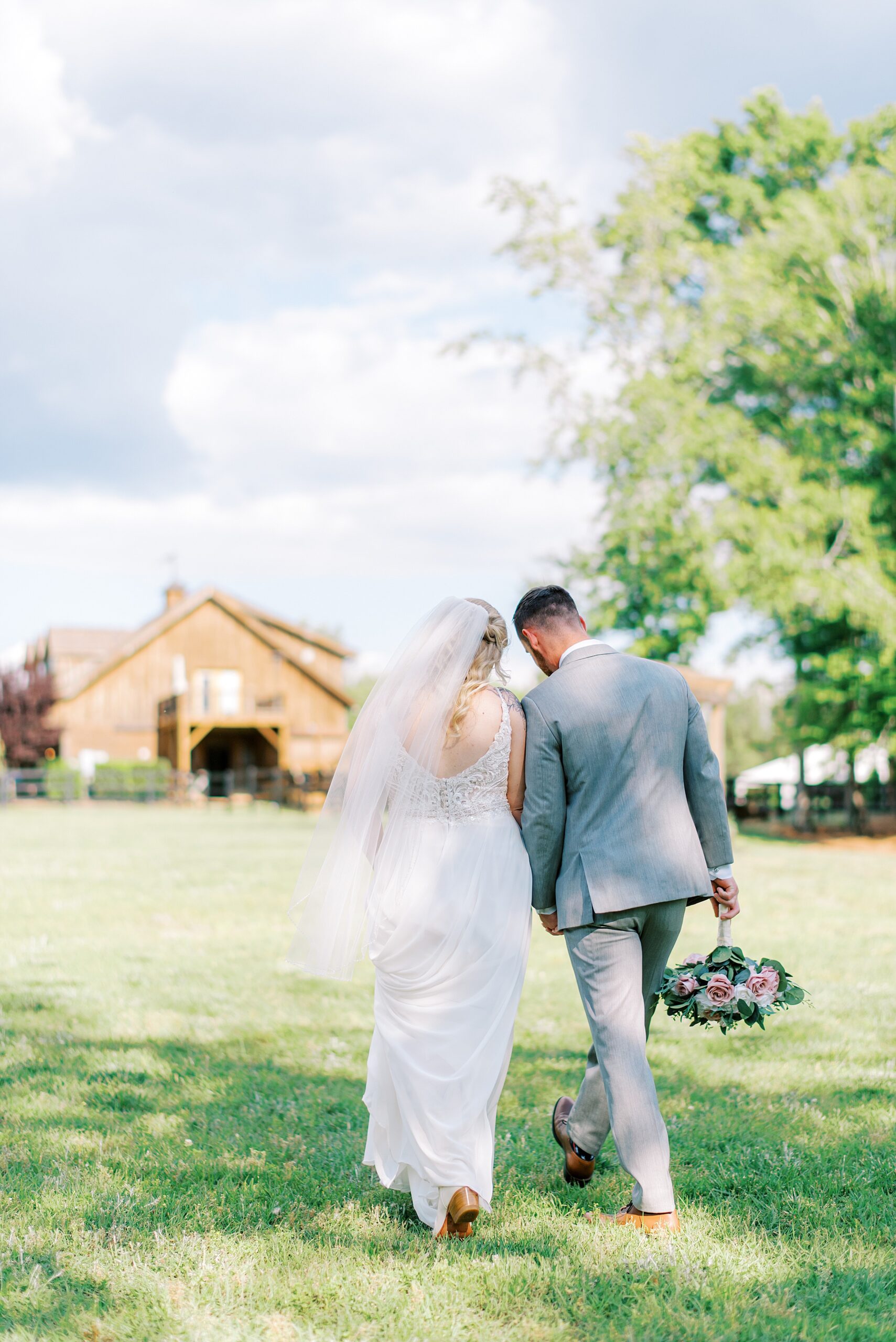 Newlyweds in a grey suit and lace dress hold hands while walking through a large field to a barn at a farm wedding venues charlotte nc